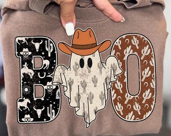 May include: A brown t-shirt with a Halloween-themed graphic. The word "BOO" is spelled out with a cowboy ghost in the center. The letters are decorated with cacti, skulls, and other western-themed images.