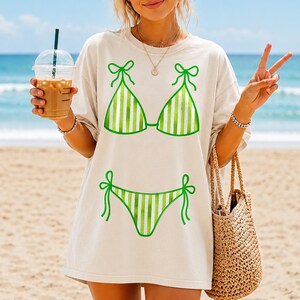 May include: Beige oversized t-shirt with a green and white striped bikini graphic. The design shows a bikini top and bottom with bow details. The image is taken on a beach, with a person holding a drink and a woven tote bag.