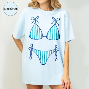 May include: Pale blue t-shirt with a bikini design. The bikini top and briefs are outlined in dark blue and have vertical stripes in shades of blue and white. The shirt has short sleeves and a loose fit. The word "chambray" is in a circle in the upper left corner.