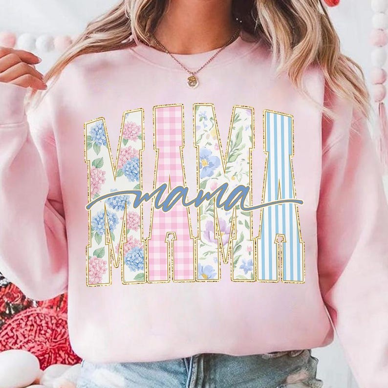 May include: Pink sweatshirt with the word "MAMA" in large, decorative letters. The letters are filled with floral, gingham, and striped patterns. The word "mama" is written in cursive below the letters.