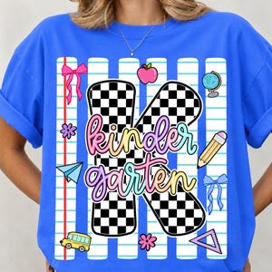 May include: A bright blue t-shirt featuring a large black and white checkered letter K with the word kindergarten written in a colorful gradient script. The design includes school-themed icons like a pencil, apple, globe, bus, and bows.