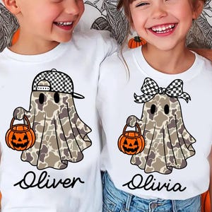 以下が含まれることがあります： ハロウィーンのゴーストデザインの白いTシャツ。1つのゴーストはチェック柄の帽子、もう1つはチェック柄のリボンを着用しています。各ゴーストはオレンジ色のカボチャのバケツを持っています。「オリバー」と「オリビア」という名前が下に書かれています。