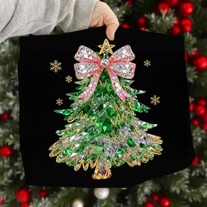 Könnte beinhalten: Schwarzes quadratisches Kissen mit einem Weihnachtsbaum-Design. Der Baum besteht aus grünen und silbernen Kristallen, mit einer rosa und Kristall Schleife oben. Goldene Schneeflocken und ein Stern akzentuieren das Design. Das Kissen wird von einer Person gehalten.