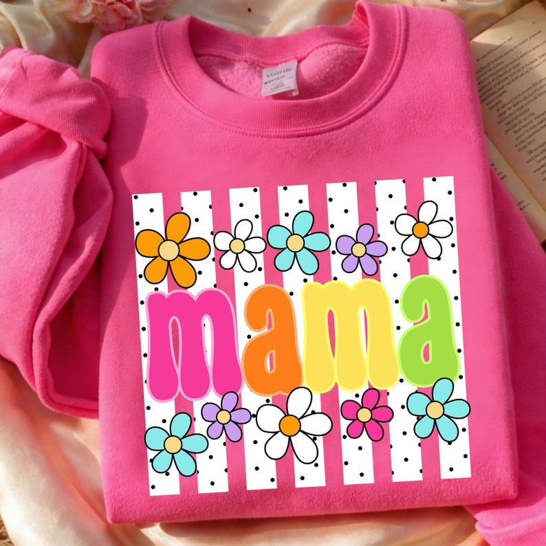 May include: A bright pink sweatshirt with a floral design and the word "mama" in colorful letters. The design includes white stripes, black polka dots, and various colorful flowers. The sweatshirt is made of a soft material.