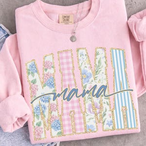May include: A light pink sweatshirt with the word "MAMA" in a bold, decorative font. The letters are filled with floral, gingham, and striped patterns. A silver necklace with a pendant is visible.