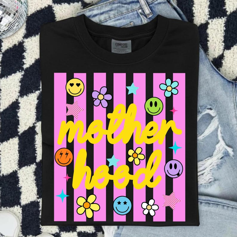 Może przedstawiać: Czarny t-shirt z r&oacute;żowym wzorem w paski. Koszulka zawiera napis "motherhood" w ż&oacute;łtym skrypcie, a także emotikony, gwiazdki i grafiki kwiatowe. Zabawny i kolorowy design.
