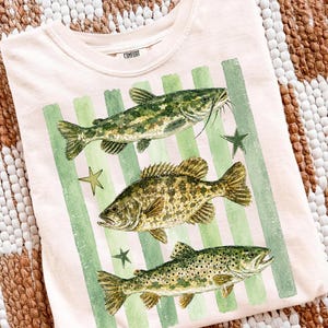 May include: A light pink t-shirt featuring three detailed fish illustrations in shades of green and brown, set against green vertical stripes. The shirt also has a small tag that says "COMFORT".