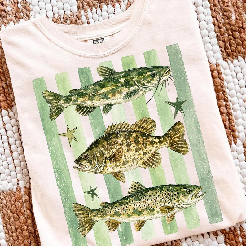 May include: A light pink t-shirt featuring three detailed fish illustrations in shades of green and brown, set against green vertical stripes. The shirt also has a small tag that says "COMFORT".