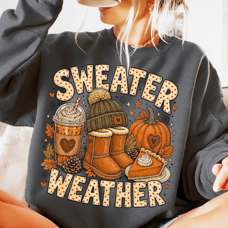 May include: A dark gray crewneck sweatshirt featuring a graphic print with the text "SWEATER WEATHER" in a dotted font. The design includes a coffee cup, a knit beanie, tan boots, a pumpkin, a slice of pumpkin pie, and autumn leaves.