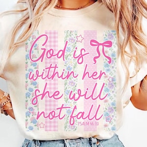 K&ouml;nnte beinhalten: Cremefarbenes T-Shirt mit rosa Blumen- und Vichy-Design. Das Shirt zeigt den Text "God is within her she will not fall" in rosa Schrift, zusammen mit dem Bibelvers Psalm 46:10. Das Design enth&auml;lt Blumen- und Sternakzente.