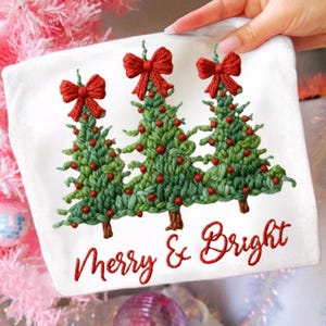 May include: White Christmas-themed hand towel featuring three green Christmas trees with red bows and ornaments. The words "Merry & Bright" are embroidered in red script below the trees. The towel is held by a hand.