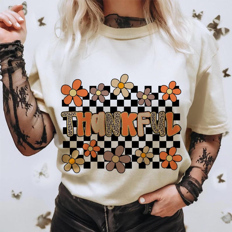May include: Cream-colored t-shirt with the word "THANKFUL" in orange and leopard print letters, set against a black and white checkered background. The shirt is decorated with orange, brown, and tan flowers.