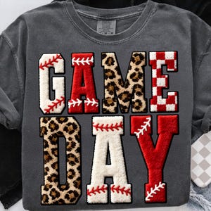 May include: Dark gray sweatshirt with the words "GAME DAY" in large, textured letters. The letters are in various styles, including baseball stitching, leopard print, and a red and white checkered pattern. The sweatshirt has a vintage look.
