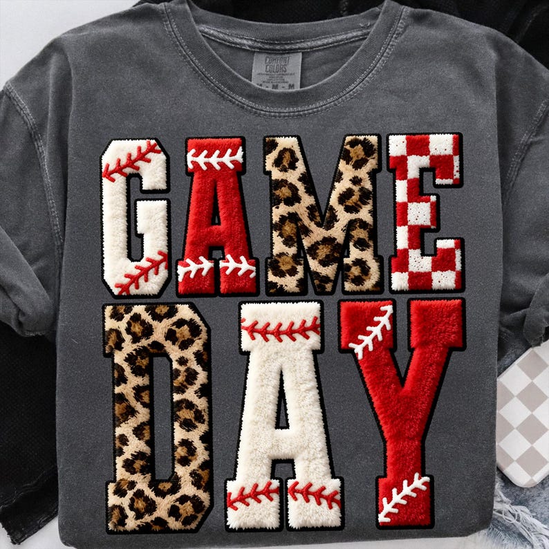 May include: Dark gray sweatshirt with the words "GAME DAY" in large, textured letters. The letters are in various styles, including baseball stitching, leopard print, and a red and white checkered pattern. The sweatshirt has a vintage look.