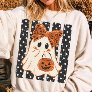 May include: A cream-colored crewneck sweatshirt featuring a central graphic of a white ghost wearing a large orange leopard-print bow. The ghost holds an orange jack-o'-lantern bucket against a background of vertical black stripes with white polka dots.