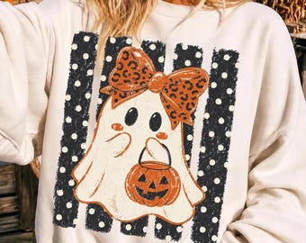 Pode incluir: Uma camisola de gola redonda cor de creme com um gráfico central de um fantasma branco a usar um grande laço cor de laranja com padrão de leopardo. O fantasma segura um balde de abóbora cor de laranja sobre riscas verticais pretas com bolas brancas.