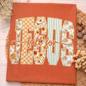 May include: A burnt orange fabric featuring the word JESUS in large block letters filled with patterns of pumpkins, autumn leaves, orange gingham, and green stripes, with the cursive text fall for overlaid in white across the centre.