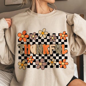 May include: Beige sweatshirt with a floral and checkered design. The word "THANKFUL" is printed in orange and leopard print. Orange, yellow, and brown flowers are arranged around the text and checkered background.