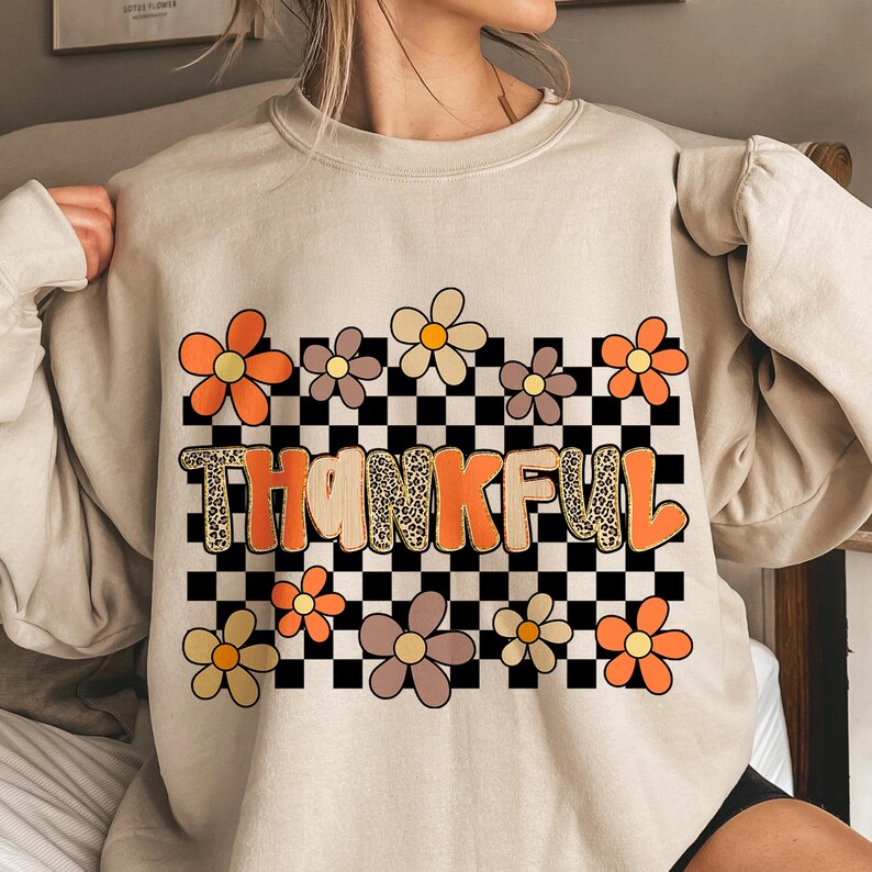 May include: Beige sweatshirt with a floral and checkered design. The word "THANKFUL" is printed in orange and leopard print. Orange, yellow, and brown flowers are arranged around the text and checkered background.