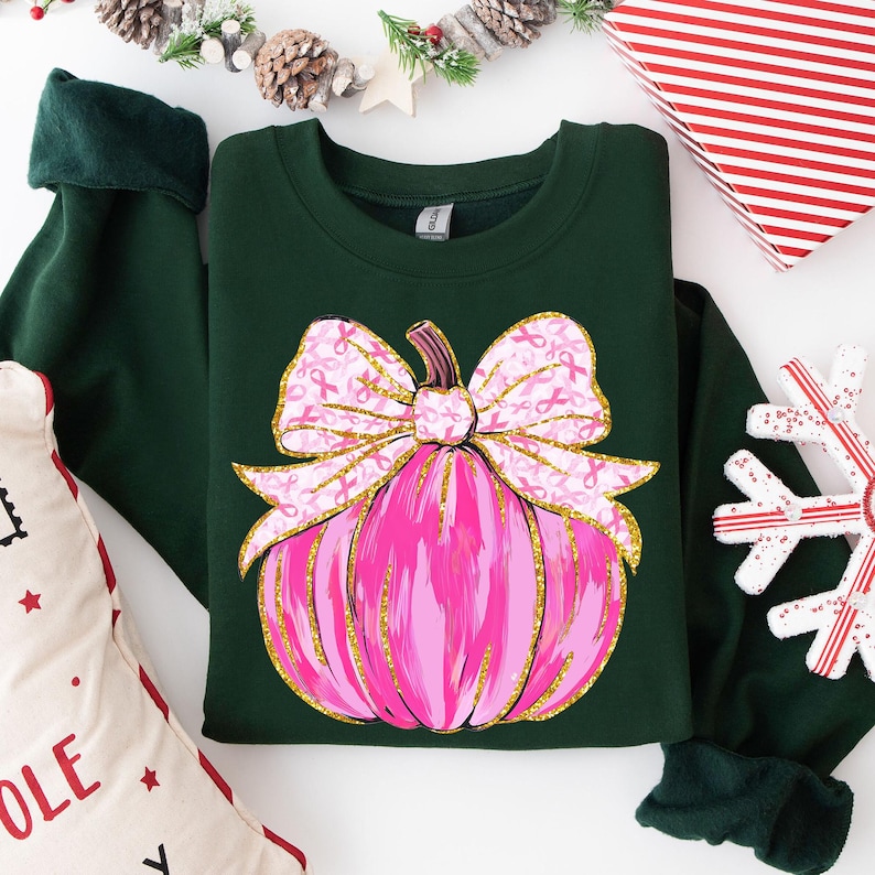 May include: A dark green sweatshirt featuring a pink pumpkin design with a gold outline and a pink bow adorned with pink ribbons. The sweatshirt is folded, with a Christmas-themed pillow and gift box in the background.