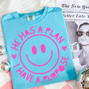 Puede incluir: Una camiseta azul claro con un dise&ntilde;o de carita sonriente rosa y el texto "HE HAS A PLAN I HAVE A PURPOSE". La camiseta tiene cuello redondo y mangas cortas. La camiseta est&aacute; hecha de un material suave.