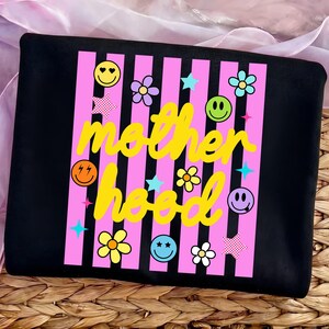 Może przedstawiać: Czarna bluza z r&oacute;żowymi pionowymi paskami i ż&oacute;łtym napisem "motherhood". Projekt zawiera emotikony, kwiaty i gwiazdki w r&oacute;żnych kolorach. Bluza jest prezentowana na wiklinowym koszu.