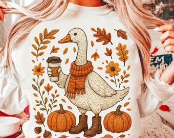 May include: White sweatshirt featuring a cartoon goose wearing a scarf and boots, holding a coffee cup. The design includes pumpkins, leaves, and flowers in shades of orange and brown, evoking an autumn theme.