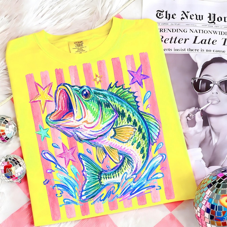 Op de afbeelding: Geel shirt met lange mouwen met een kleurrijke illustratie van een largemouth bass. Het ontwerp bevat roze strepen, sterren en waterspetters. Het shirt heeft een ronde hals en een label met "CURIOS".