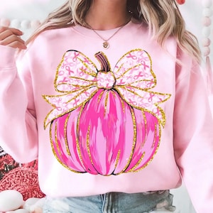 May include: Pink sweatshirt featuring a pink pumpkin design with a bow. The bow is decorated with pink ribbons and gold glitter accents. The pumpkin also has gold glitter outlines. The sweatshirt is a solid light pink color.