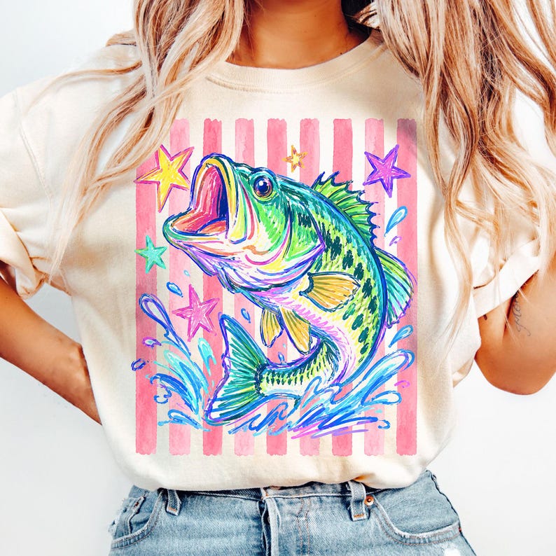 Op de afbeelding: Cr&egrave;mekleurig T-shirt met een levendige illustratie in aquarel-stijl van een largemouth bass die uit het water springt. Het ontwerp bevat roze en witte strepen, kleurrijke sterren en waterspetters. De vis is groen, blauw en geel.