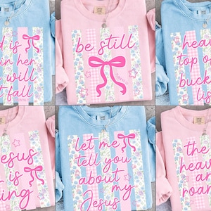 K&ouml;nnte beinhalten: Eine Auswahl an pastellfarbenen Sweatshirts mit inspirierenden Zitaten. Jedes Shirt hat ein einzigartiges Design mit Text, B&auml;ndern und floralen Mustern. Die Zitate beinhalten S&auml;tze wie "Gott ist in ihr" und "Jesus ist K&ouml;nig."