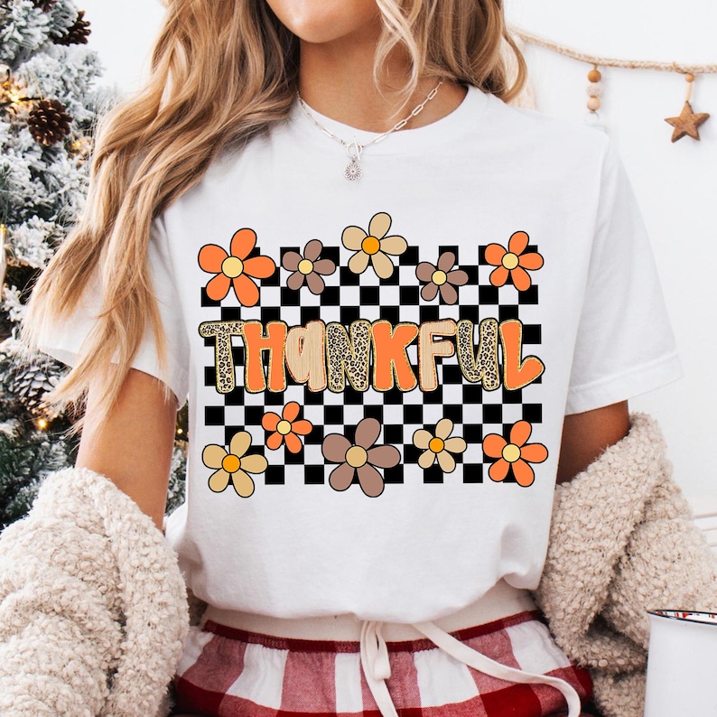 May include: White t-shirt with the word "THANKFUL" in orange and tan letters, set against a black and white checkered background. The shirt is decorated with orange, tan, and brown flowers.