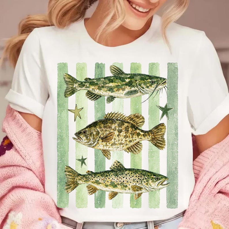 May include: White t-shirt featuring three detailed fish illustrations in shades of green and brown, set against a backdrop of green vertical stripes and star accents. The design has a vintage, outdoorsy aesthetic.
