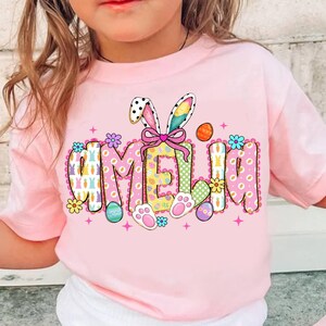 May include: A light pink t-shirt featuring the name "AMELIA" in a colorful, Easter-themed design. The design includes bunny ears, Easter eggs, flowers, and paw prints. The shirt is a short-sleeved tee.