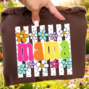 May include: Brown sweatshirt with the word "mama" in colorful, retro-style lettering. The word is set against a white background with black polka dots and vertical stripes, and surrounded by colorful flower graphics.