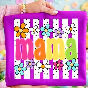 May include: A bright purple fabric item featuring a central graphic with the word mama in colorful, rounded letters. The design includes a background of vertical white stripes with black polka dots and various multi-colored flower illustrations.