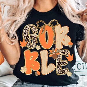 May include: Black t-shirt with the word "GOBBLE" in orange, leopard print, and wood-grain lettering. The design includes fall leaves, acorns, and a turkey. A bow is above the "O".