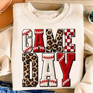 May include: Cream-colored sweatshirt with the words "GAME DAY" in a bold, layered design. The letters feature baseball stitching, leopard print, and red and white checkered patterns, perfect for sports fans.