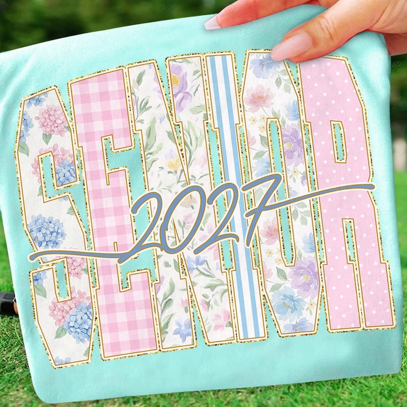 May include: A light blue t-shirt with the word "SENIOR" in a collegiate font, with the year "2027" in the center. The letters are filled with floral, gingham, and polka dot patterns, outlined in gold.