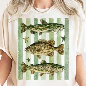 May include: Cream-colored t-shirt featuring a graphic of three fish: a catfish, a bass, and a trout, set against a backdrop of green and white vertical stripes. The design includes green star accents. The shirt has short sleeves.