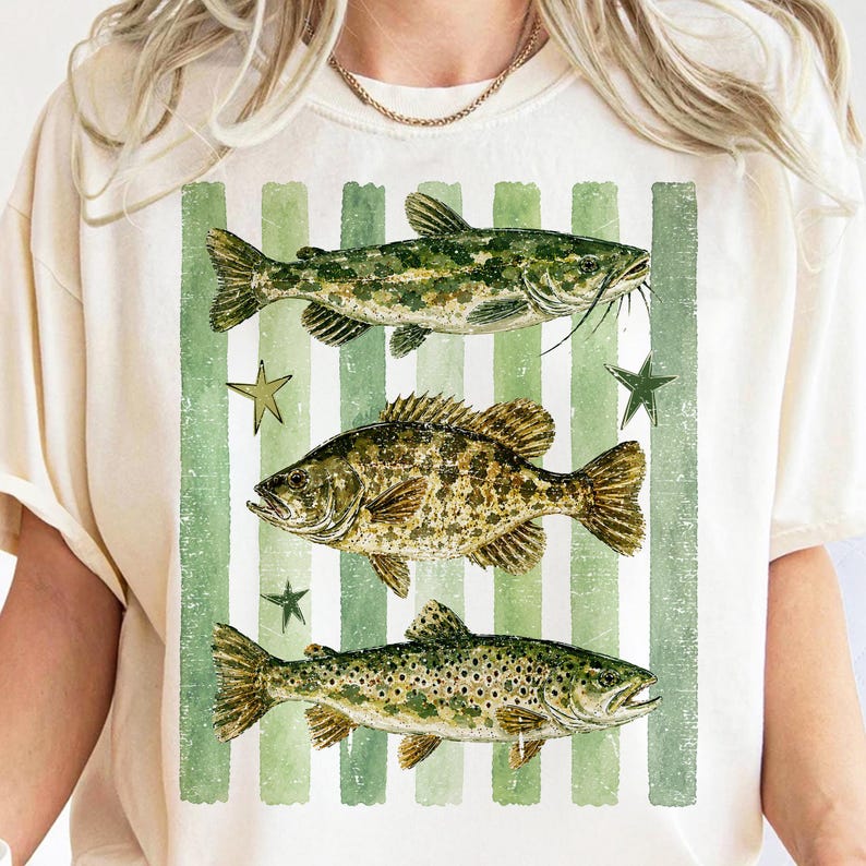May include: Cream-colored t-shirt featuring a graphic of three fish: a catfish, a bass, and a trout, set against a backdrop of green and white vertical stripes. The design includes green star accents. The shirt has short sleeves.