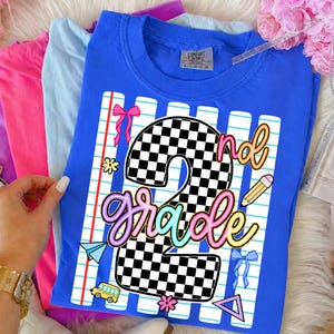 May include: A blue short-sleeve t-shirt featuring a graphic of a large checkered number 2 and the text 2nd grade in colorful script. The design includes a lined paper background, a yellow pencil, a yellow school bus, and pink bows.