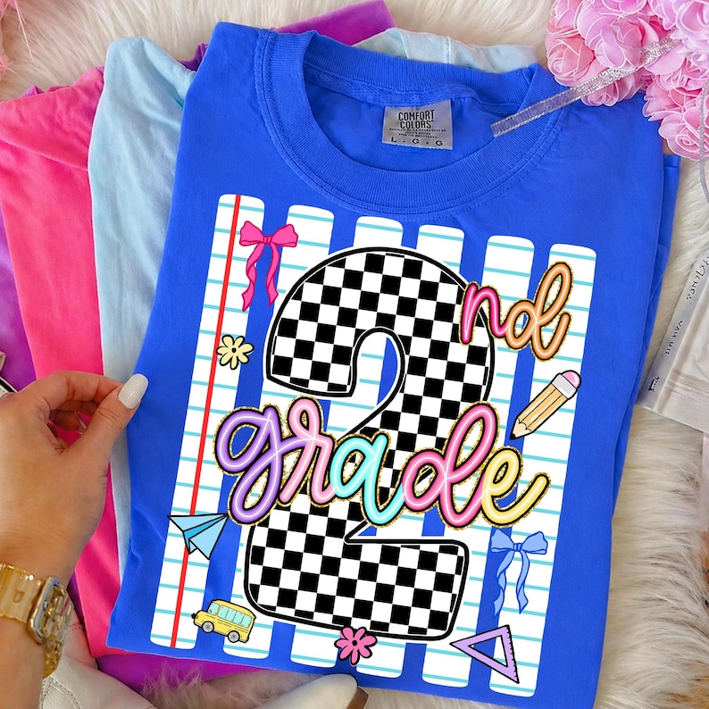 May include: A blue short-sleeve t-shirt featuring a graphic of a large checkered number 2 and the text 2nd grade in colorful script. The design includes a lined paper background, a yellow pencil, a yellow school bus, and pink bows.