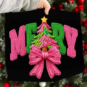 May include: Black fabric with a knitted design that says "MERRY" in pink and green. A pink Christmas tree and bow are in the center, with a gold star on top of the tree. The design is held up by a person.