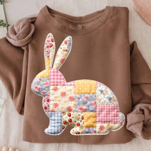 Puede incluir: Una sudadera marrón con un diseño de conejo de patchwork. El conejo está hecho de varios cuadrados de tela estampada, incluyendo patrones florales, cuadros y lunares. La sudadera tiene mangas largas y cuello redondo.