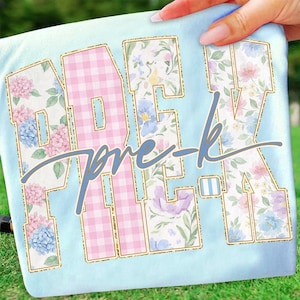 May include: Pale blue t-shirt featuring the text "PRE-K" in large, floral-patterned letters with gold glitter outlines. The letter "E" is pink gingham. The words "pre-k" are written in cursive in a light blue colour.