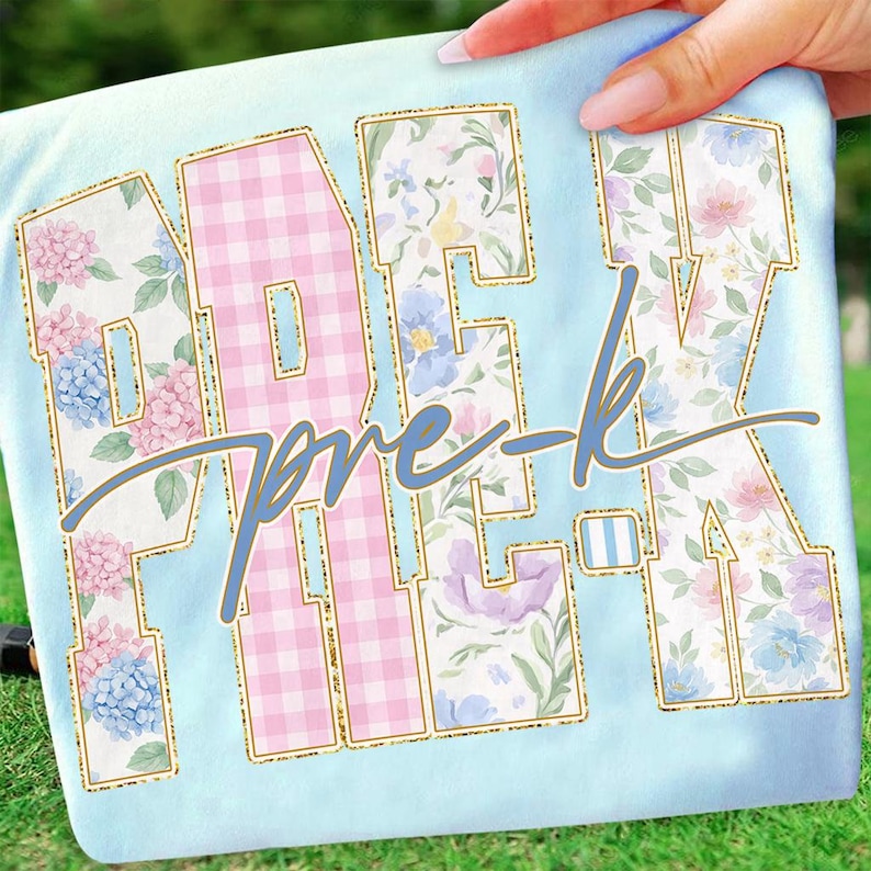 May include: Pale blue t-shirt featuring the text "PRE-K" in large, floral-patterned letters with gold glitter outlines. The letter "E" is pink gingham. The words "pre-k" are written in cursive in a light blue colour.