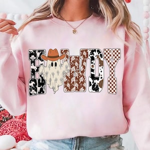 K&ouml;nnte beinhalten: Ein hellrosa Sweatshirt mit dem Wort "HALLOWDY" in einem Western-Design. Die Buchstaben weisen Muster wie Kuhdruck, Kakteen und ein Schachbrettmuster auf. Ein Geist mit Cowboyhut befindet sich in der Mitte.