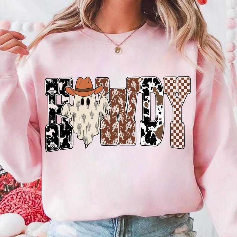 K&ouml;nnte beinhalten: Ein hellrosa Sweatshirt mit dem Wort "HALLOWDY" in einem Western-Design. Die Buchstaben weisen Muster wie Kuhdruck, Kakteen und ein Schachbrettmuster auf. Ein Geist mit Cowboyhut befindet sich in der Mitte.