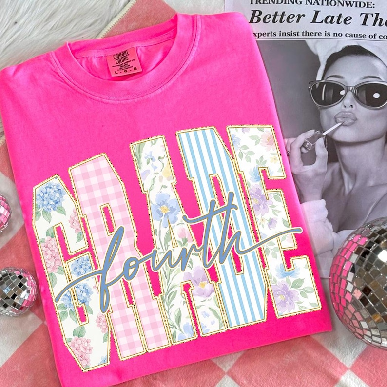May include: A vibrant pink t-shirt featuring the text "GRADE FOURTH". The letters are adorned with floral, gingham, and blue striped patterns. The shirt is a Comfort Colors brand.